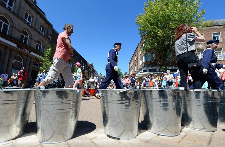A sunny day with a blue sky in a town centre. We see a line of shiny, steel buckets in the foreground with people walking past and a crowd in the background